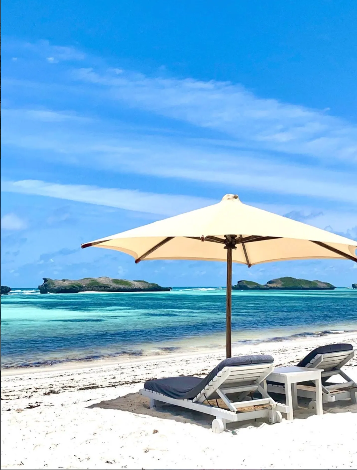 A serene tropical beach scene with white sand, turquoise ocean, lounge chairs under a beige umbrella, and distant rocky islands under a clear blue sky.