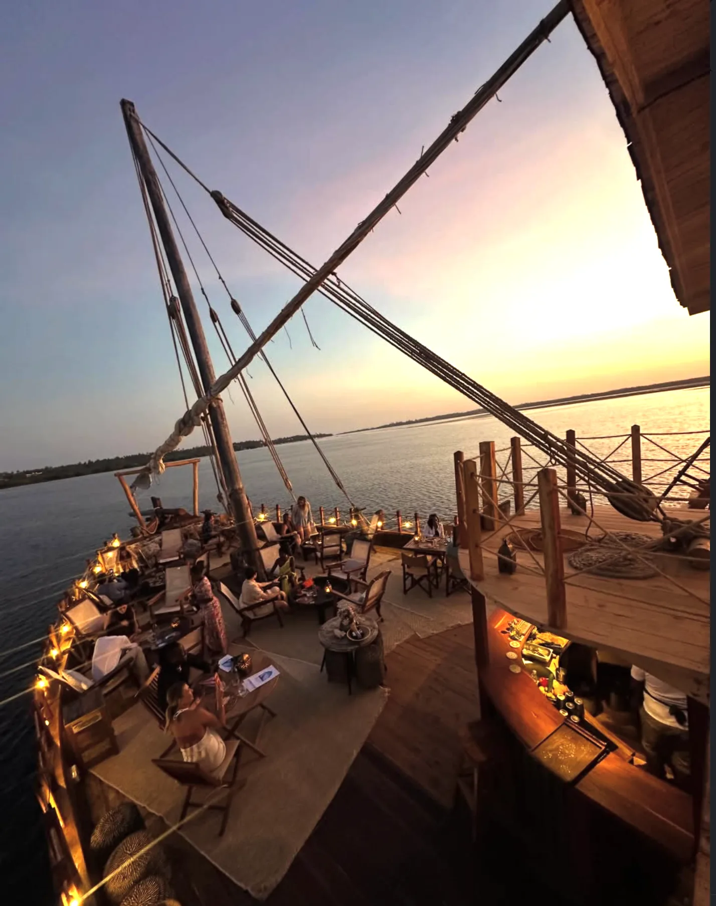 A lively outdoor bar on a traditional dhow boat at sunset with people relaxing by the water.