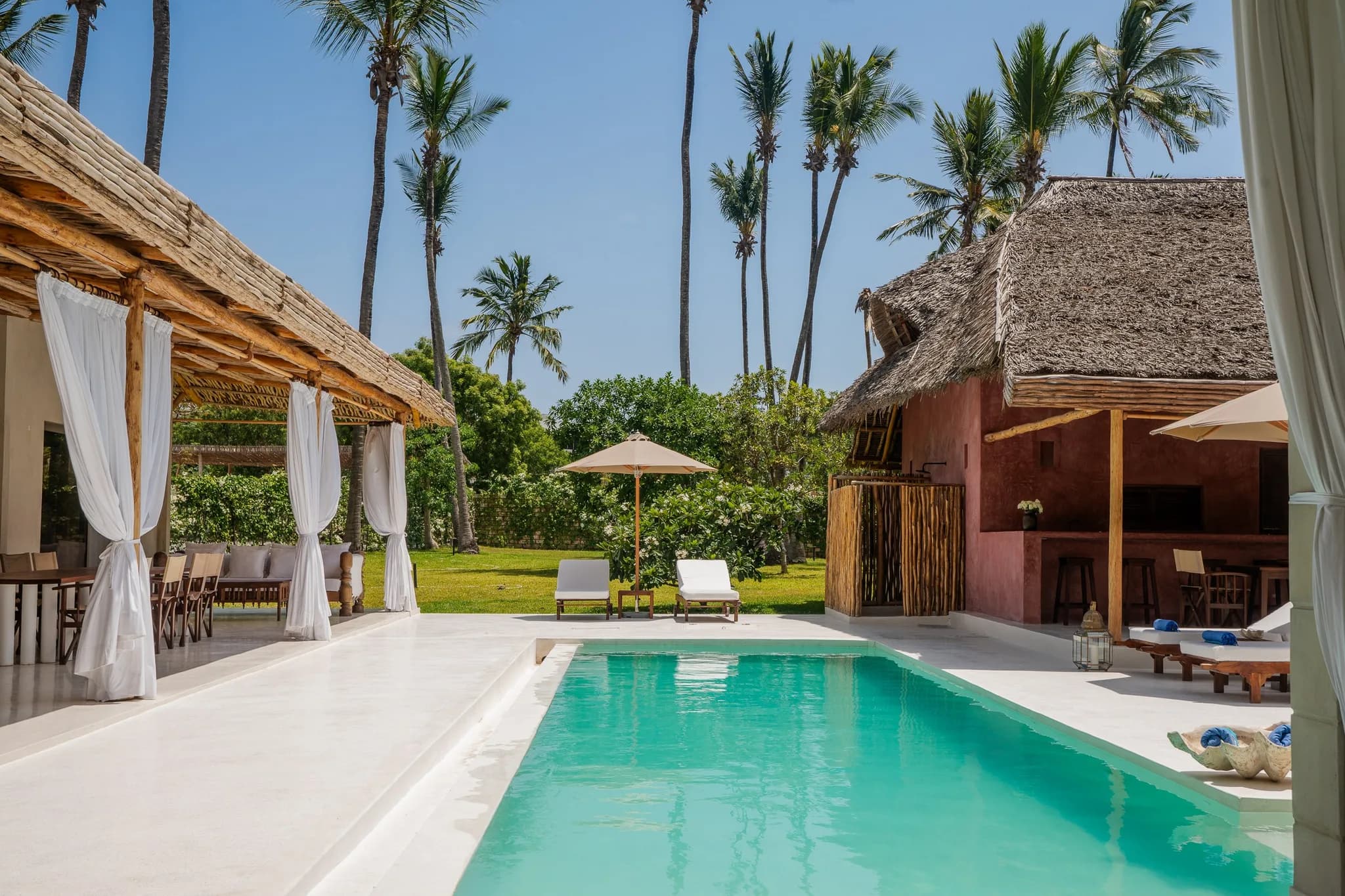 A wide-angle shot of a tropical villa complex with palm trees, pool, outdoor seating, and lush greenery under blue skies.