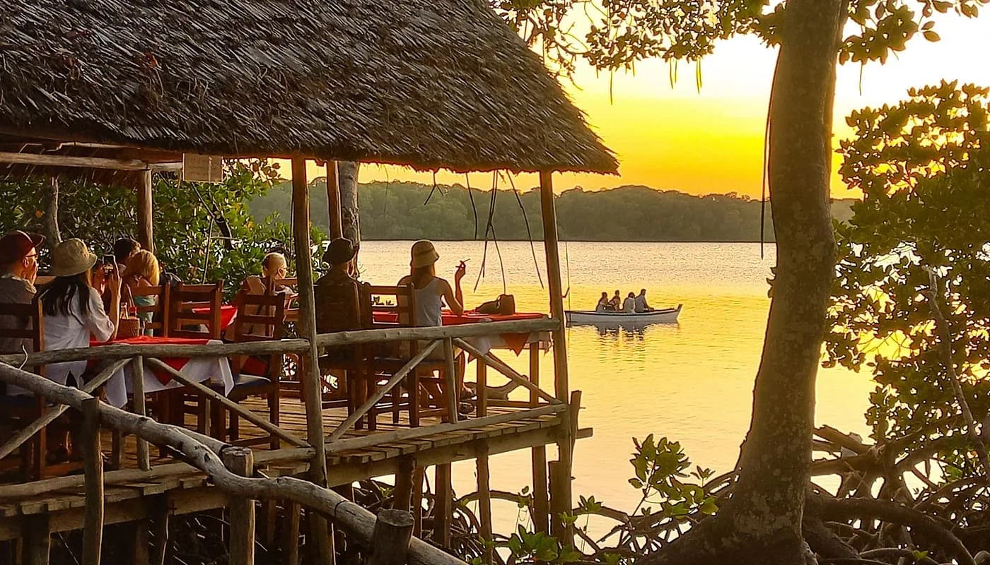Tourists dine at an open-air waterfront restaurant on a wooden deck overlooking a serene lake with a canoe and lush mangroves at sunset.