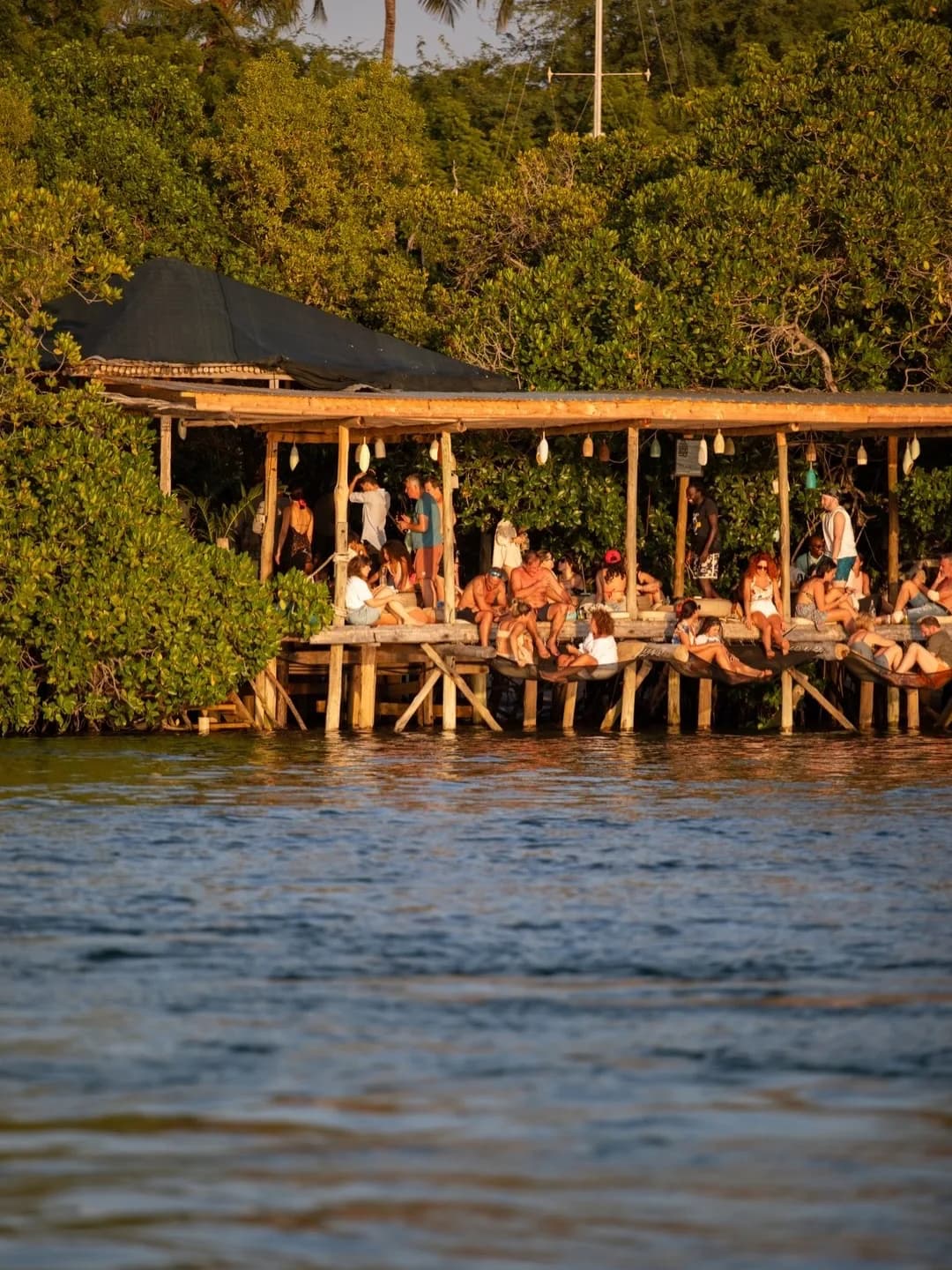 A lively group of people relaxes on a wooden beach bar structure surrounded by mangroves over calm water at sunset, evoking a tropical, carefree vibe.