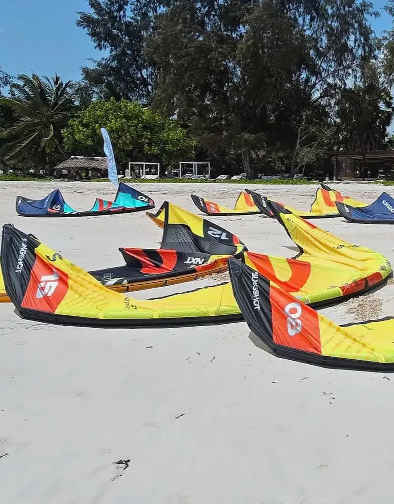 Colorful kitesurfing kites of various designs lie deflated on a sunny tropical beach surrounded by palm trees and oceanfront huts, evoking a relaxed vacation mood.