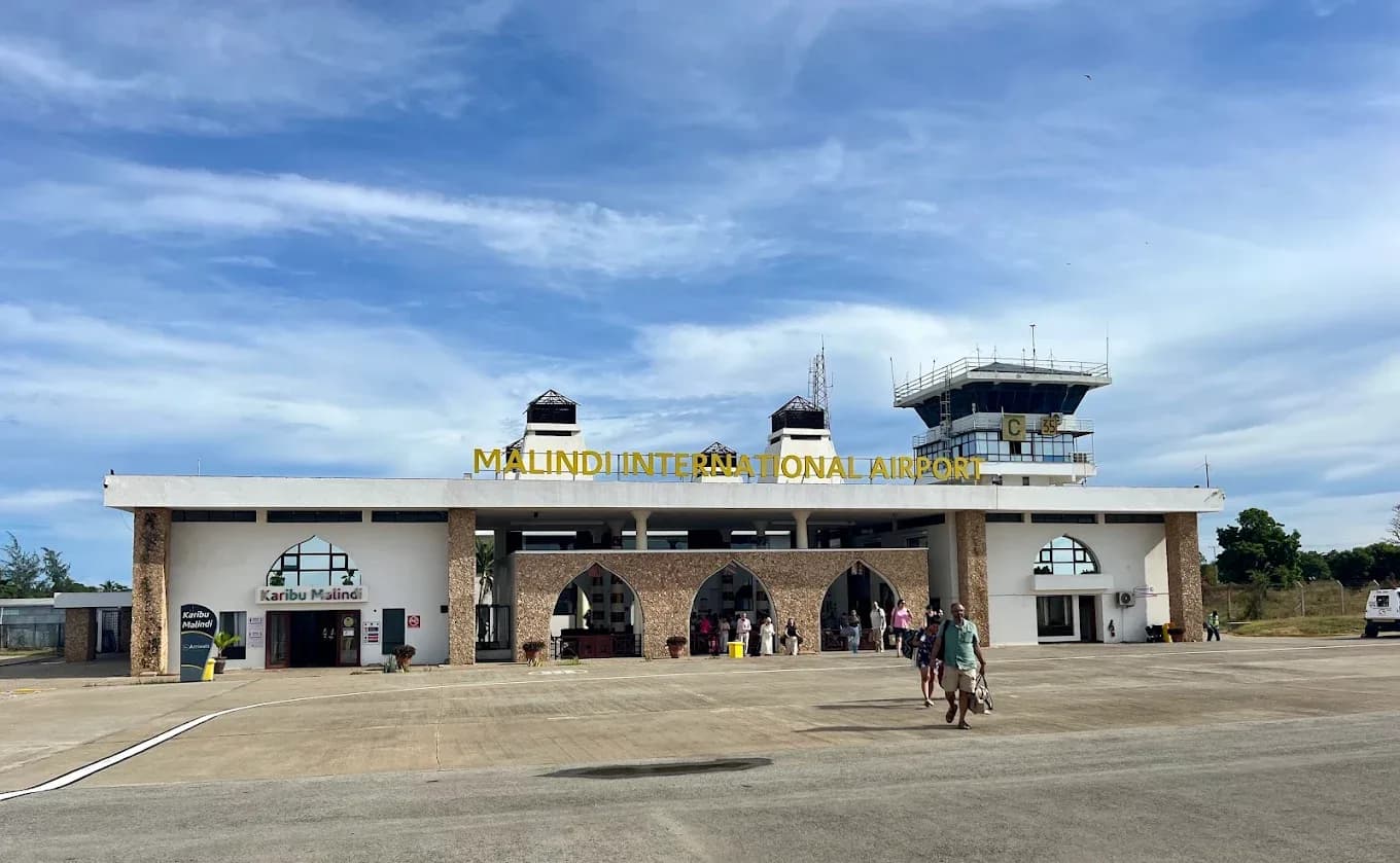 The exterior of Malindi International Airport features its modern terminal building and control tower under a partly cloudy sky with people walking nearby.